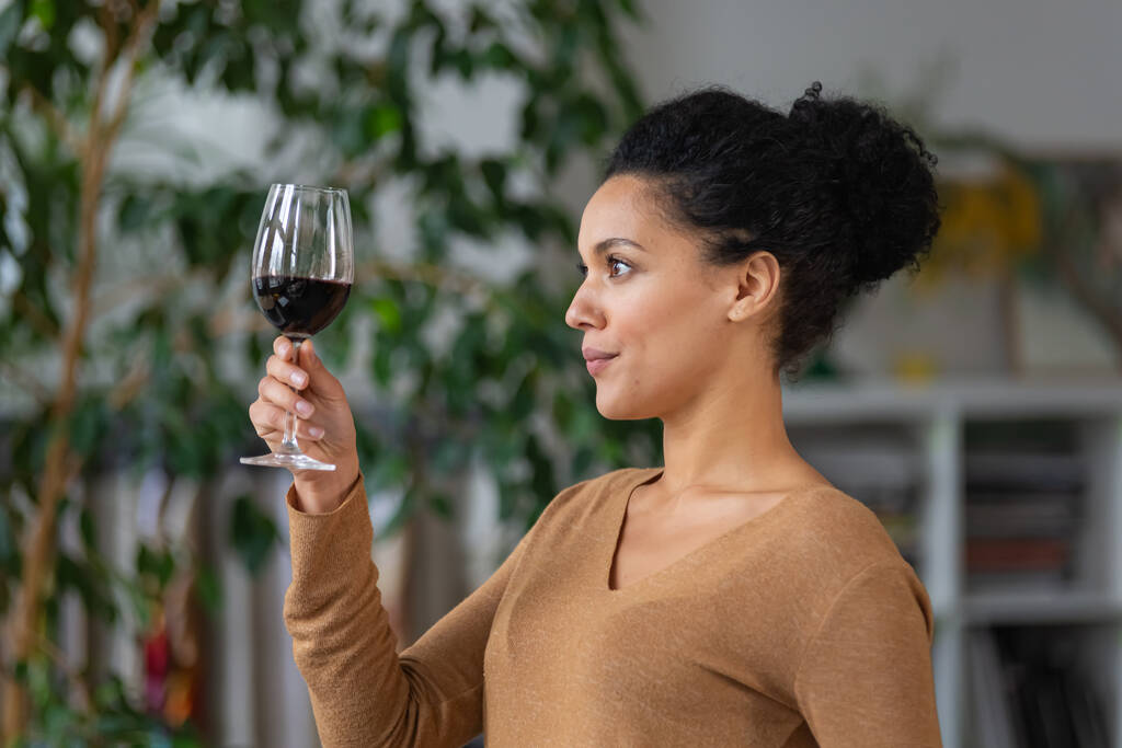 Portret van een jonge Afro-Amerikaanse vrouw kijkend naar een glas rode wijn. Schattig gemengd ras vrouwelijke poseren tegen wazig achtergrond van lichte kamer met groene planten. Sluiten.. - Foto, afbeelding