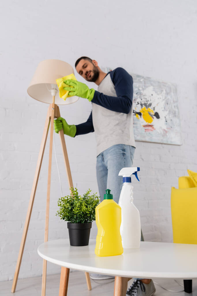 Niski kąt widzenia detergentów na stole w pobliżu rozmytego człowieka czyszczenia lampy podłogowej  - Zdjęcie, obraz