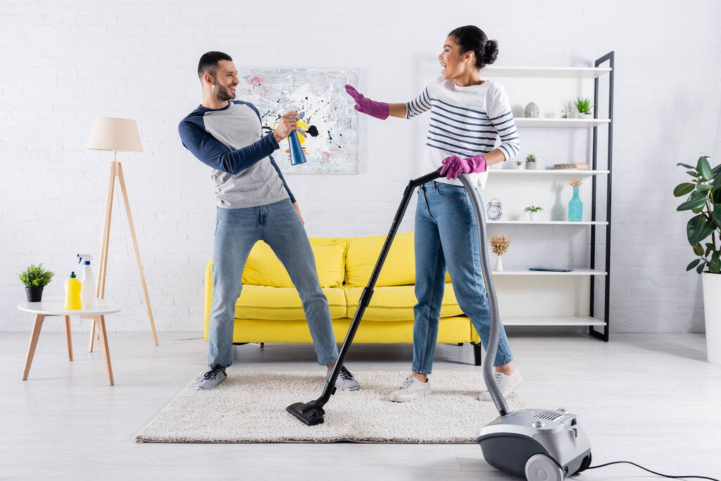 Side view of cheerful man holding detergent near african american girlfriend with vacuum cleaner  - Photo, Image