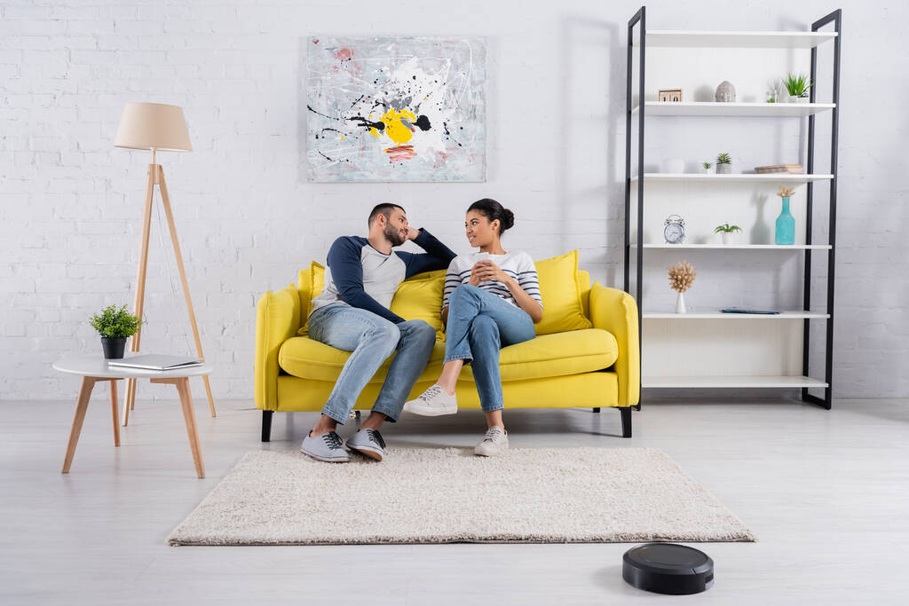 Jeune couple interracial avec smartphone parlant près d'aspirateur robotique  - Photo, image