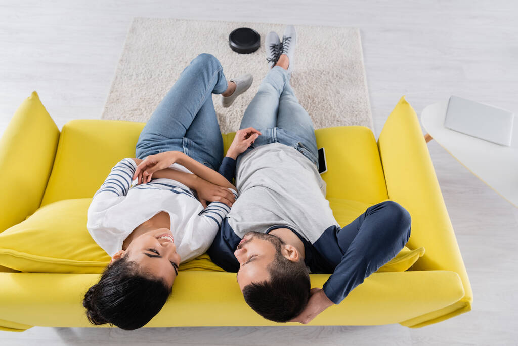 Вид сверху улыбающейся многонациональной пары, сидящей на диване рядом с размытой роботизированной уборщицей на ковре  - Фото, изображение