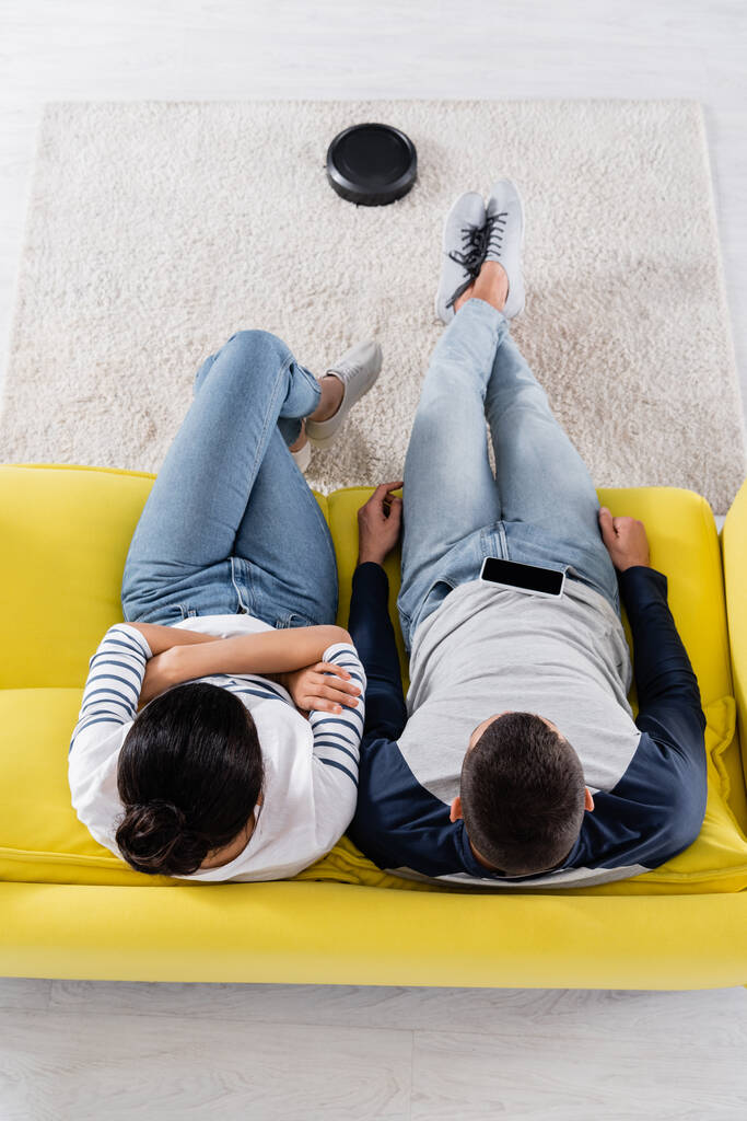 Vista aérea de la pareja multiétnica sentada en el sofá cerca de la aspiradora robótica en la alfombra  - Foto, Imagen