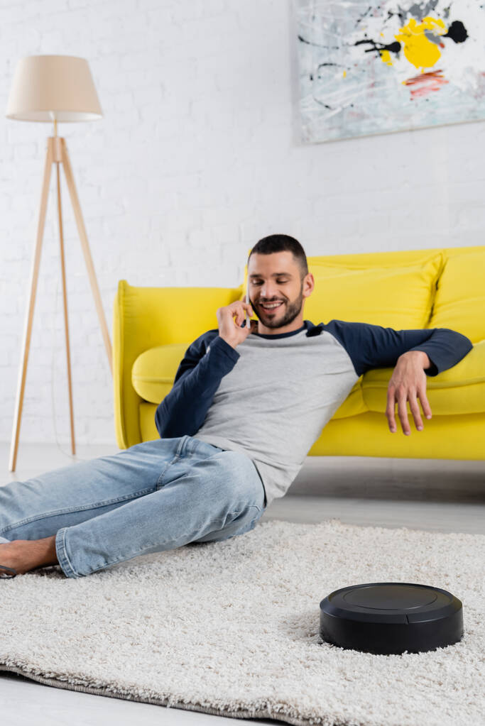 Роботизований пилосос на килимі біля усміхненого чоловіка, що розмовляє на смартфоні на розмитому фоні
  - Фото, зображення
