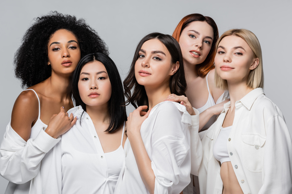 Asijské a kudrnaté africké americké modely v bílých košilích pózují se ženami izolovanými na šedé - Fotografie, Obrázek