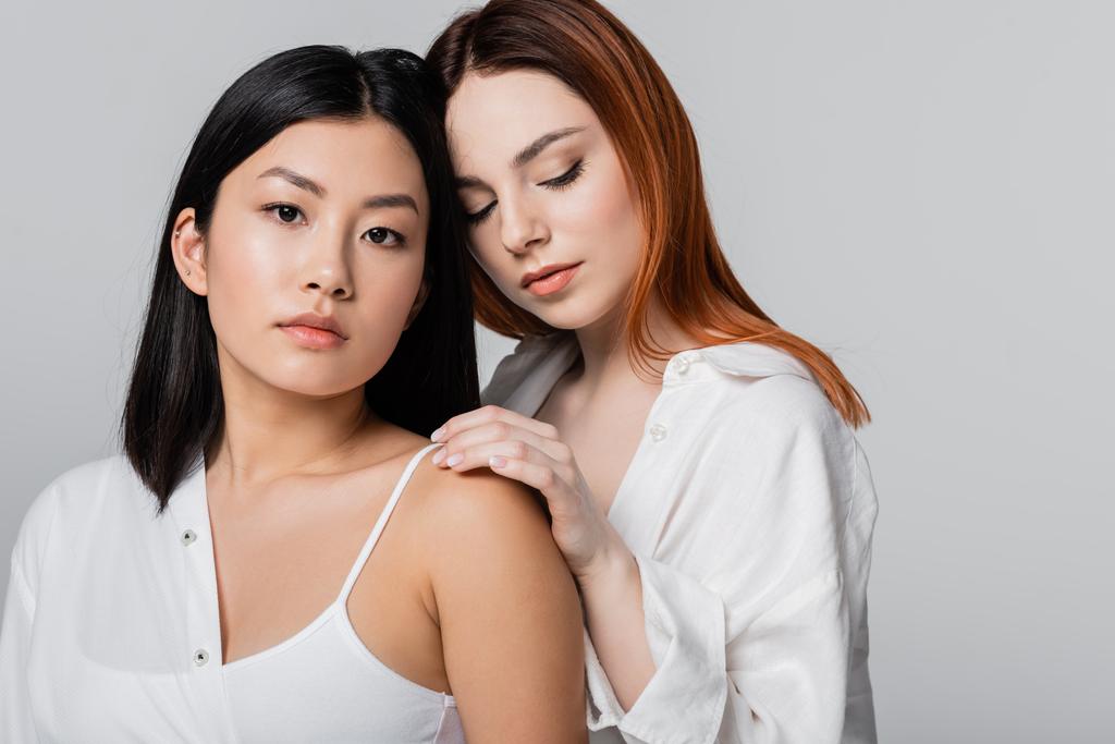 брюнетка азиатская женщина и рыжая модель позируют вместе изолированы на сером - Фото, изображение