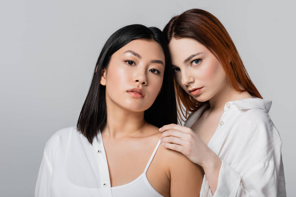 jong roodharige model poseren met brunette aziatische vrouw geïsoleerd op grijs - Foto, afbeelding