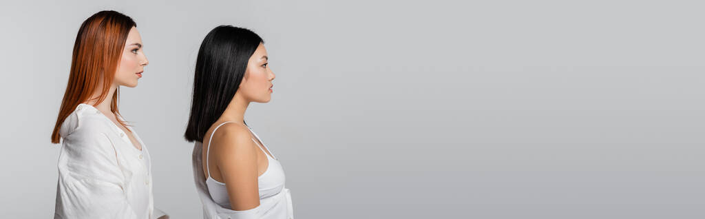 vista lateral da morena asiática mulher e ruiva modelo posando isolado no cinza, banner - Foto, Imagem