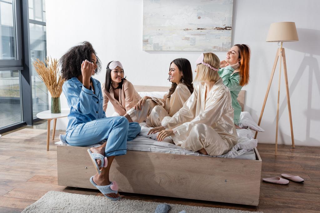 grupa wesoły międzyrasowy przyjaciele w piżama siedzi na łóżko podczas piżama party  - Zdjęcie, obraz