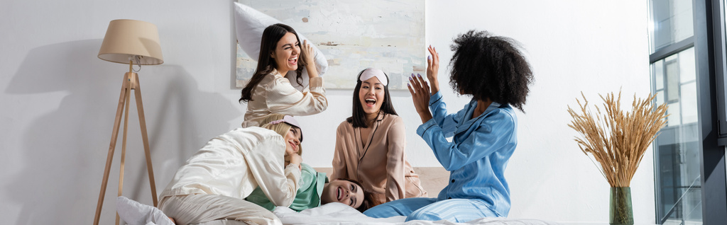 group of cheerful interracial friends in pajamas having pillow fight during slumber party, banner - Photo, Image