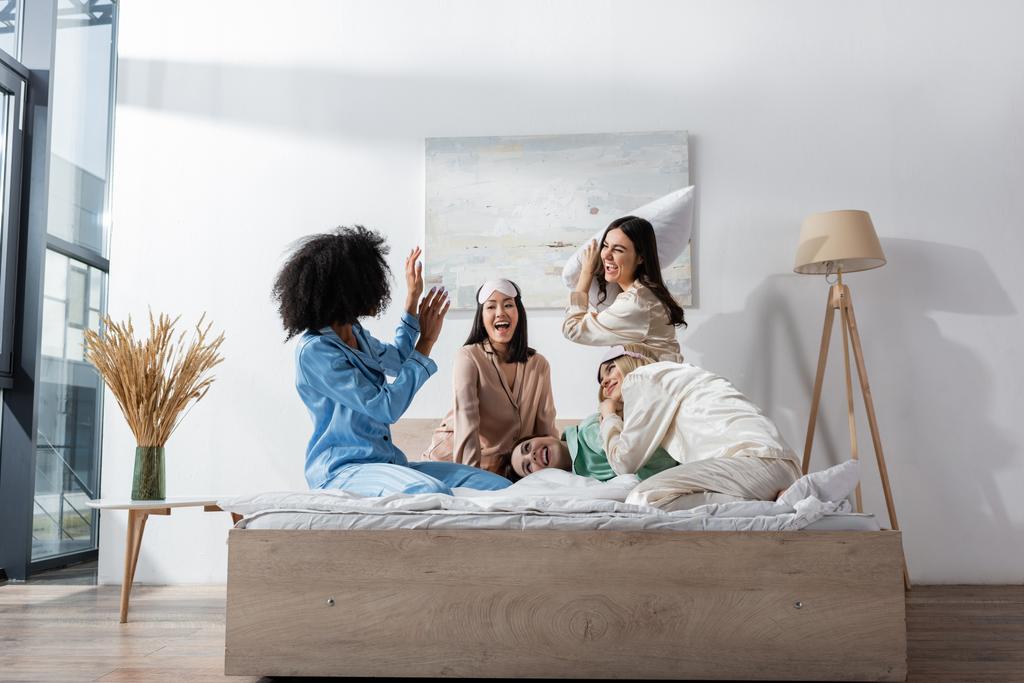group of amazed and happy interracial friends in pajamas having pillow fight during slumber party  - Photo, Image