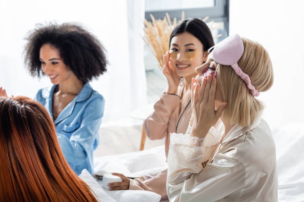 blonde woman applying eye patches near blurred interracial friends during slumber party  - Photo, Image