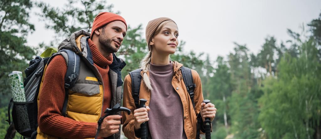couple with backpacks holding hiking sticks and looking away in forest, banner - Photo, Image