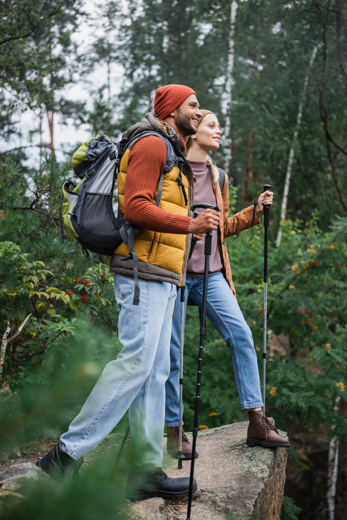 森の中で彼氏の近くを歩くハイキング棒を持つ幸せな女性の完全な長さ  - 写真・画像