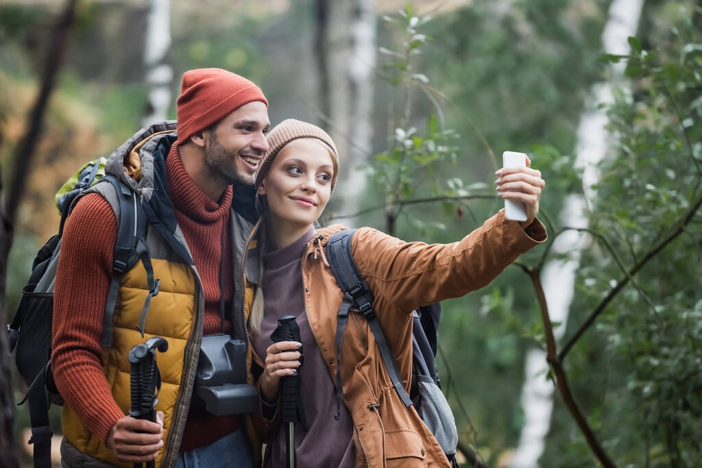 cheerful couple taking selfie with hiking sticks in forest  - Fotoğraf, Görsel