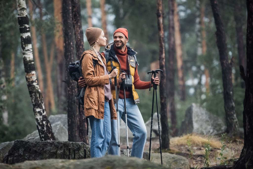 Fröhliches Paar mit Hüten hält Wanderstöcke in der Hand, während es sich im Wald umarmt - Foto, Bild