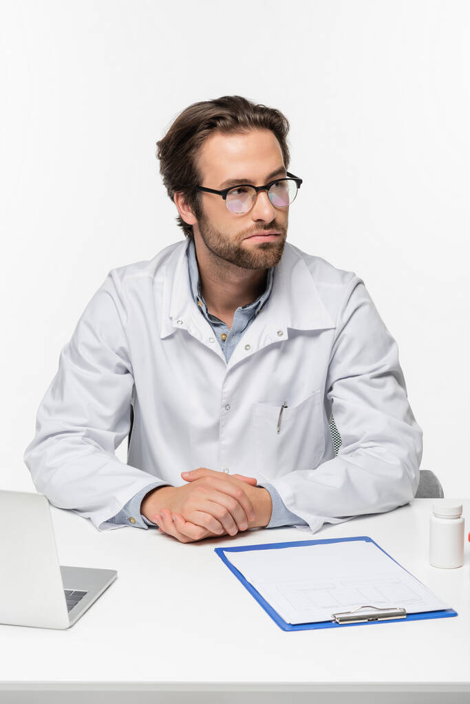 γιατρός κάθεται κοντά πρόχειρο, φορητό υπολογιστή και δοχείο με χάπια που απομονώνονται σε λευκό, ιατρική έννοια κάνναβης - Φωτογραφία, εικόνα