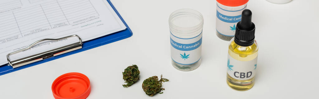 白い机の上のクリップボードの近くに油と乾燥した医療大麻のコンテナ - 写真・画像