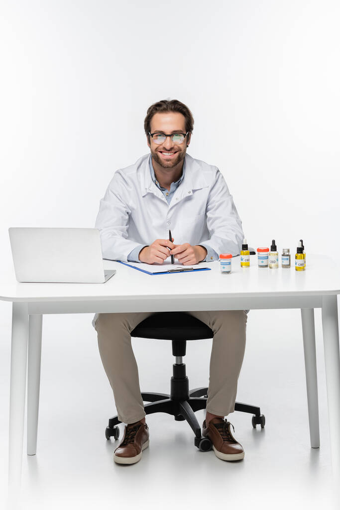 medico felice seduto alla scrivania con laptop e contenitori con cannabis medica su bianco - Foto, immagini