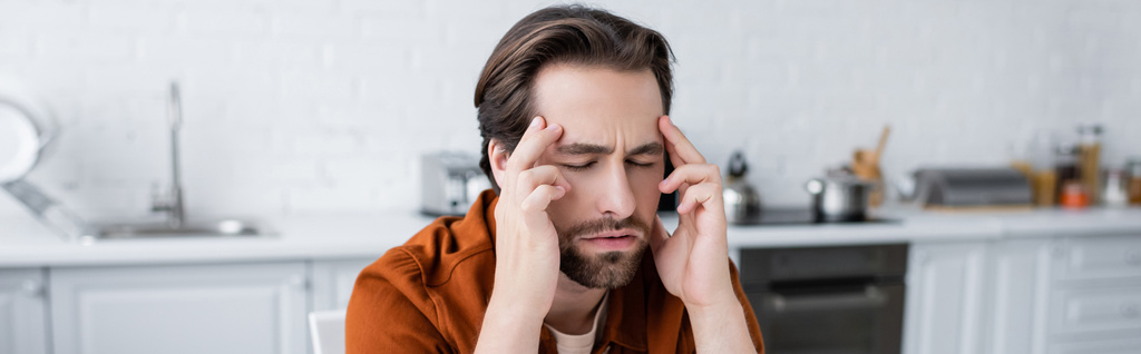 homem tocando cabeça enquanto sofre de dor de cabeça com os olhos fechados na cozinha, banner - Foto, Imagem