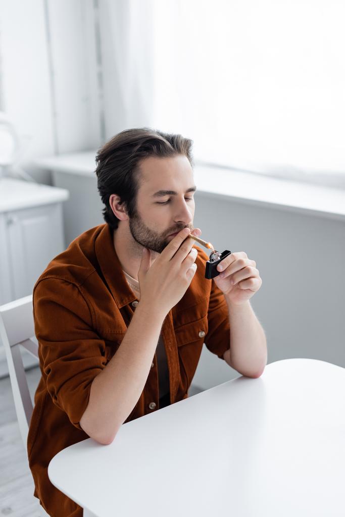 Hombre sosteniendo más ligero mientras fuma cannabis medicinal en casa  - Foto, imagen