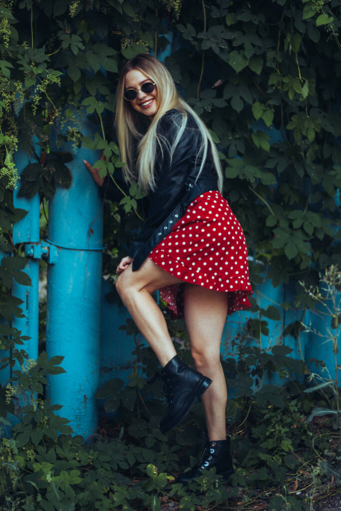retrato urbano rua de jovem bela mulher elegante em vestido vermelho e jaqueta de couro preto usando óculos de sol. fica perto de uma cerca azul entrelaçada com plantas verdes - Foto, Imagem