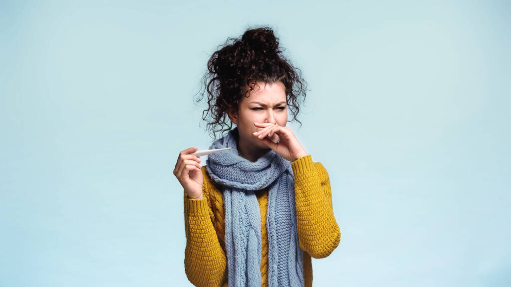 donna malata che tiene il termometro e pulisce il naso con la mano isolata sul blu - Foto, immagini
