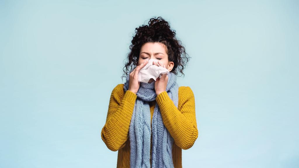 άρρωστη γυναίκα με ζεστό πουλόβερ και μαντήλι φτάρνισμα σε χαρτοπετσέτα απομονωμένη σε μπλε - Φωτογραφία, εικόνα