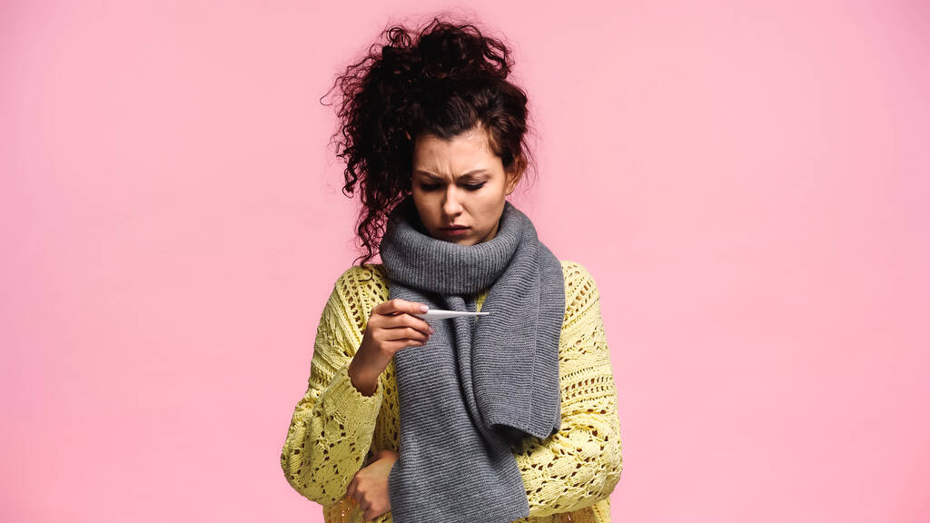 Unzufriedene Frau in Schal und Pullover blickt auf Thermometer isoliert auf rosa - Foto, Bild