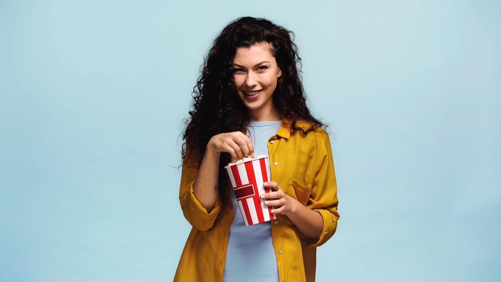 veselá žena jíst popcorn a usmívat se kamera izolované na modré - Fotografie, Obrázek