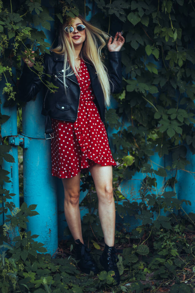 retrato urbano de la calle de la joven hermosa mujer con estilo en vestido rojo y chaqueta de cuero negro con gafas de sol. se encuentra cerca de una cerca azul entrelazada con plantas verdes - Foto, Imagen