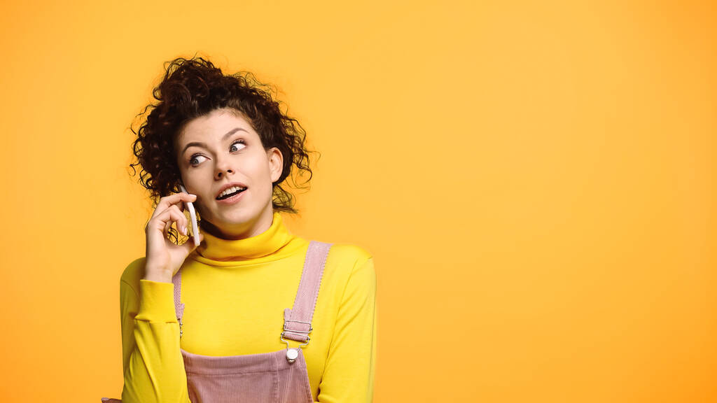 amazed woman in yellow turtleneck talking on smartphone isolated on orange - Photo, Image