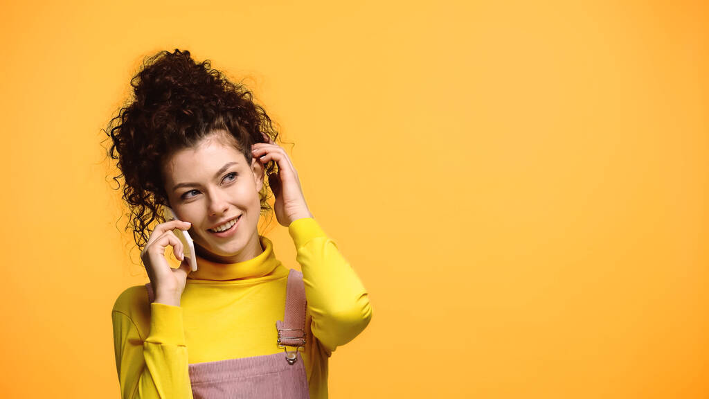 mulher sorridente ajustando o cabelo durante a conversa no celular isolado na laranja - Foto, Imagem