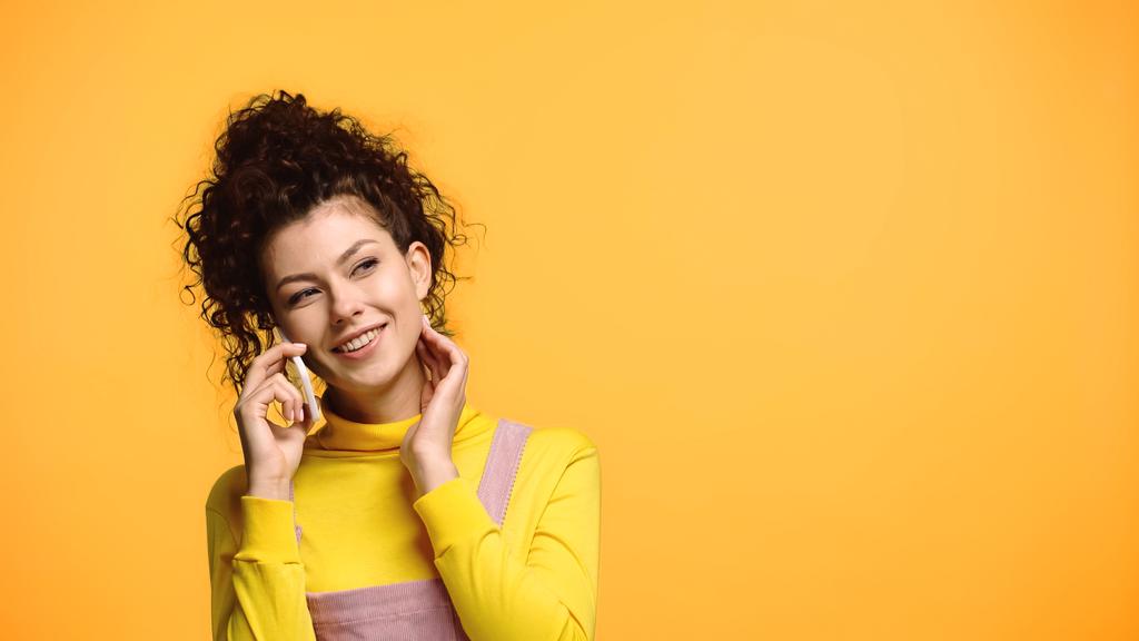 pleased woman holding hand near neck while talking on cellphone isolated on orange - Photo, Image