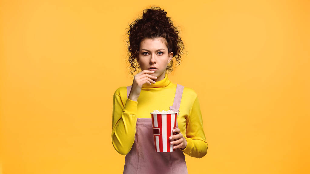 bezorgde vrouw met emmer popcorn kijkend naar camera geïsoleerd op oranje - Foto, afbeelding