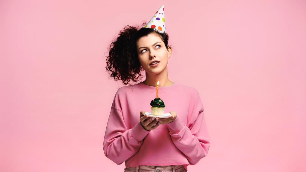 Hayalperest kadın, doğum günü pastasını pembenin üzerinde izole bir şekilde tutarken dilek tutuyor. - Fotoğraf, Görsel