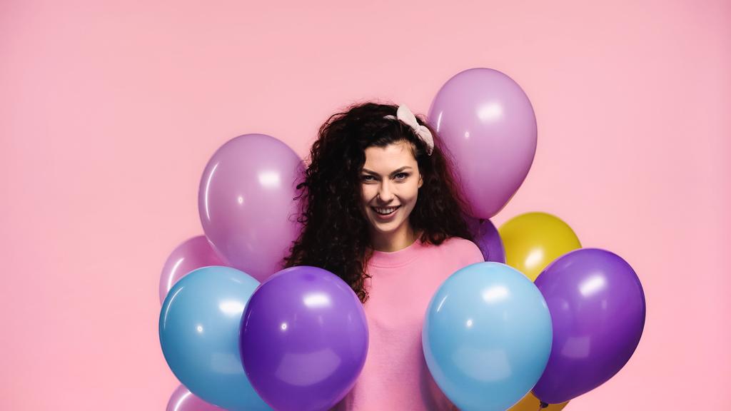 vrolijke vrouw glimlachen op camera in de buurt van veelkleurige ballonnen geïsoleerd op roze - Foto, afbeelding