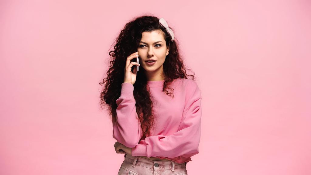morena mujer con el pelo rizado hablando en el teléfono inteligente aislado en rosa - Foto, Imagen