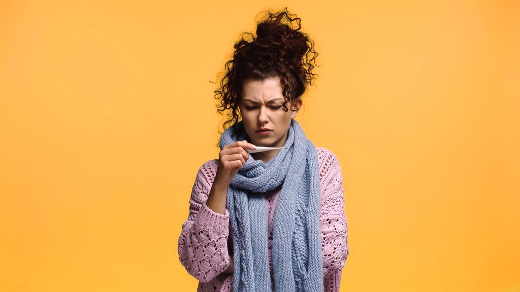 elégedetlen nő kötött pulóverben és sállal nézi hőmérő elszigetelt narancs - Fotó, kép
