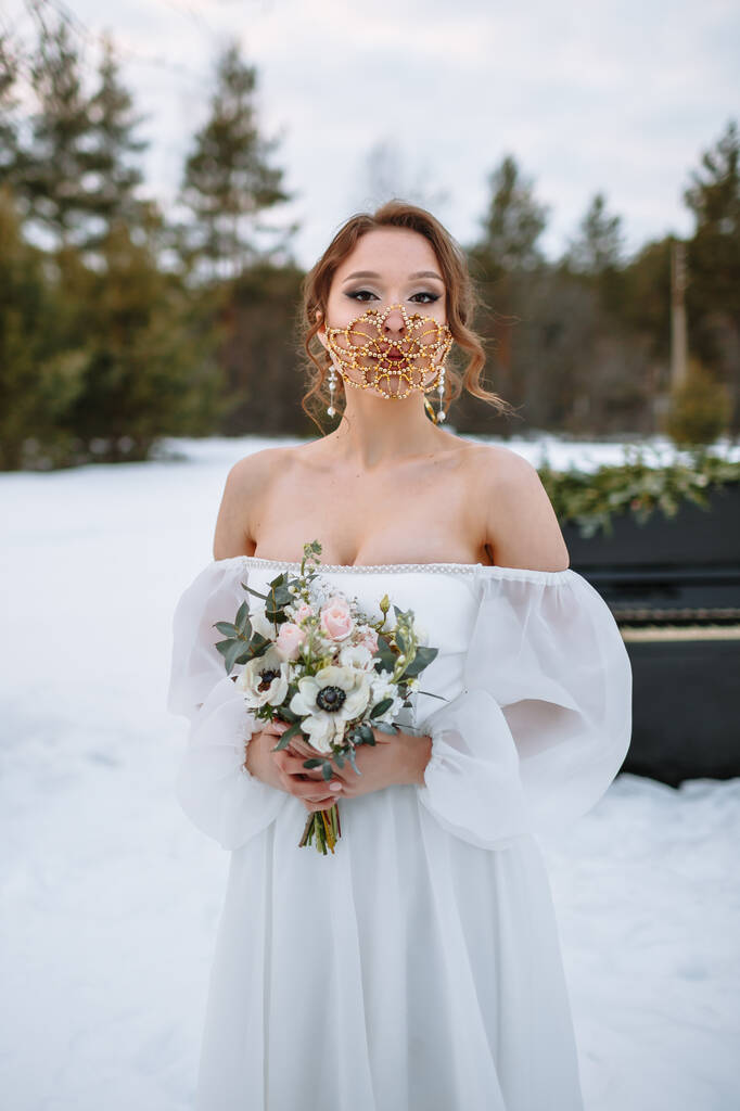 La novia está parada en un bosque cubierto de nieve. La novia lleva una fabulosa máscara protectora.. - Foto, Imagen