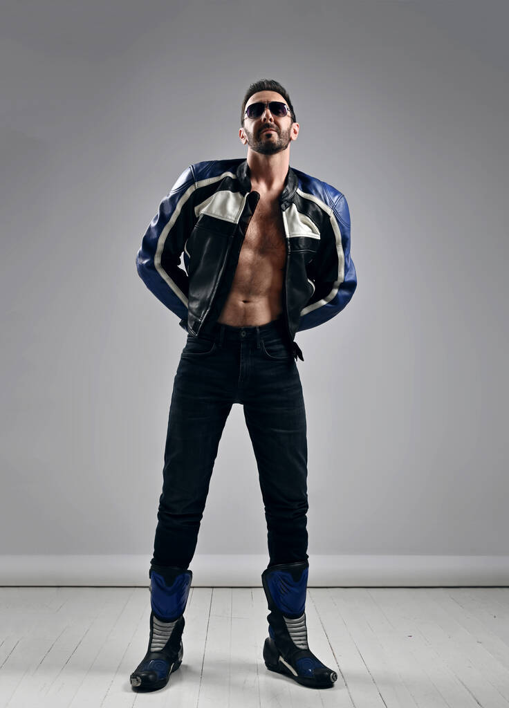 Brutal self-confident unshaved man biker in motorcycle outfit leather jacket on naked body, jeans and boots stands - Photo, Image