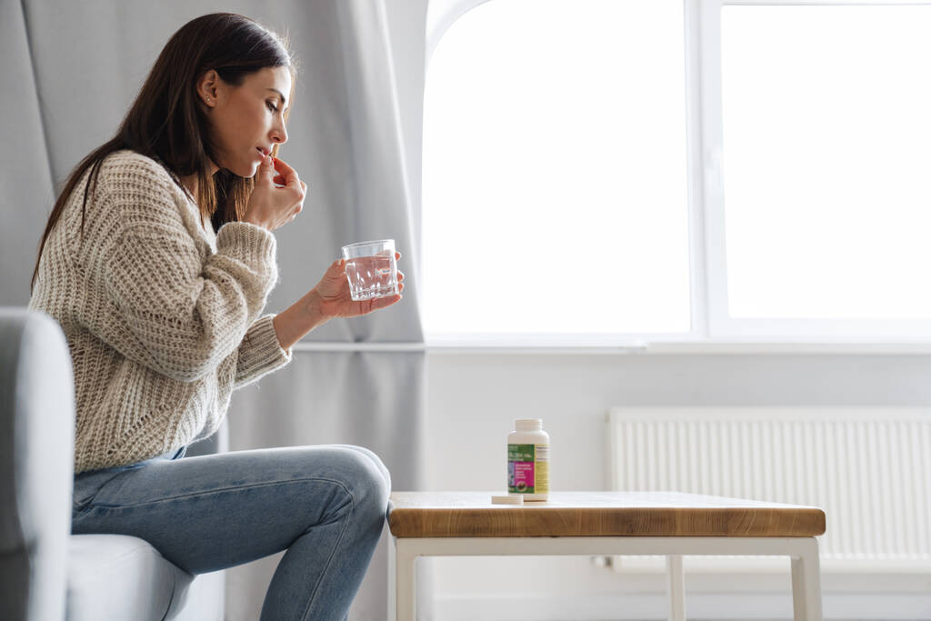 Boční pohled na ženu, jak si bere pilulku s vodou, zatímco sedí na gauči ve světlém pokoji - Fotografie, Obrázek