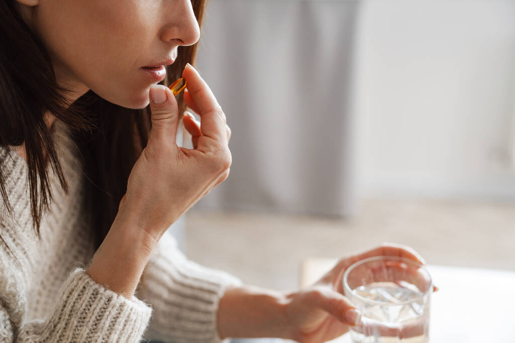 Крупный план женщины, держащей желтую таблетку во рту и держащей стакан воды, сидя в комнате - Фото, изображение