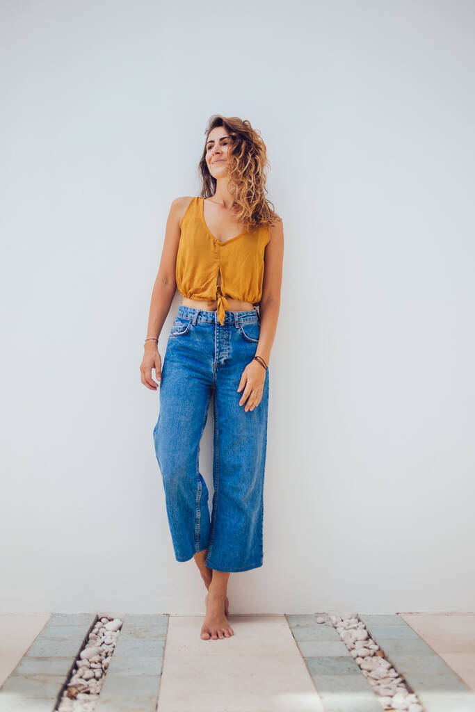 Barefoot female in yellow top and blue jeans standing on stone floor and leaning on white wall while looking away  - Photo, Image