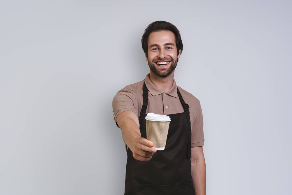 Veselý mladý muž v zástěře drží šálek kávy a usmívá se, zatímco stojí na šedém pozadí - Fotografie, Obrázek