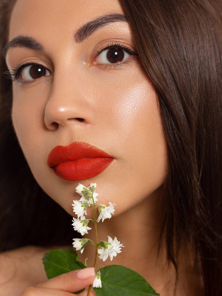 Close-up beauty of female face withevening make-up. Black arrows on the eyes and extremely long eyelashes, on full red lips matte scarlet lip color. Well-groomed skin. white flowers - Photo, Image