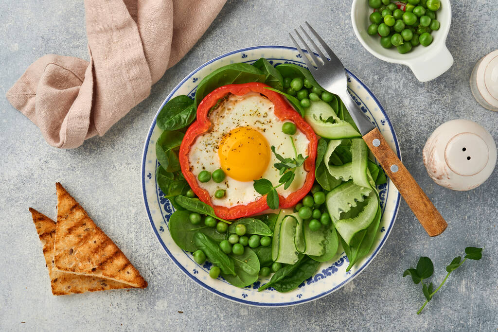 Красный перец, фаршированный яйцами, листьями шпината, зеленым горошком и микрозеленью на тарелке для завтрака на светло-сером фоне стола. Вид сверху. - Фото, изображение
