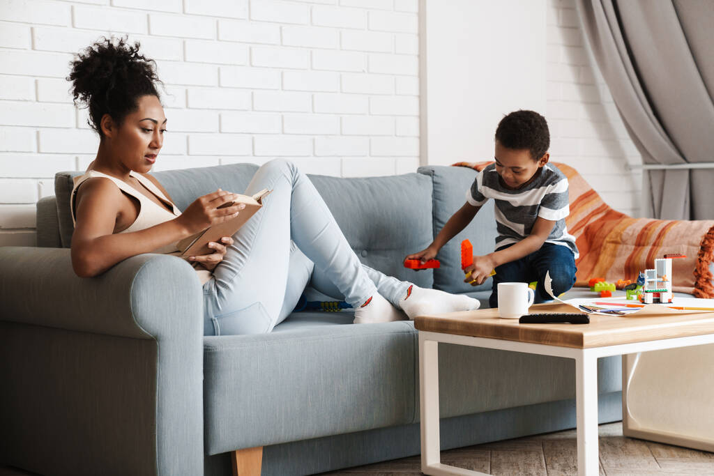 Femme noire lecture livre tandis que son fils jouer avec des jouets à la maison - Photo, image