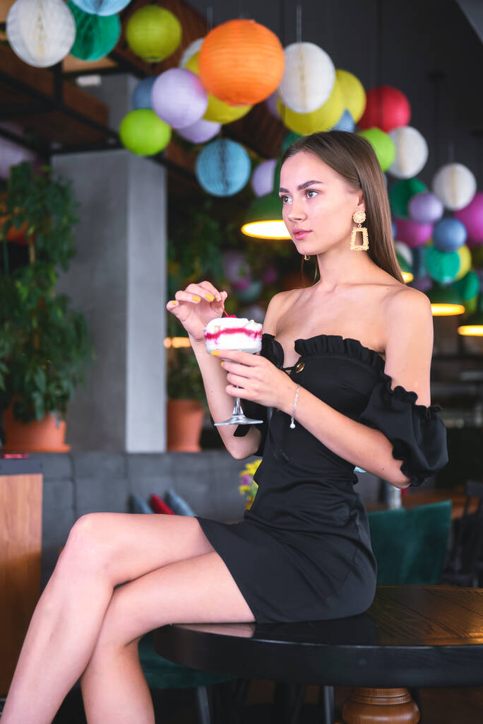 Mooie sexy brunette vrouw in restaurant of cafe met ijs tiramisu dessert. Fashion foto van jonge vrouw in zwarte jurk met kapsel en make-up, het eten van dessert. Uit eten concept. - Foto, afbeelding