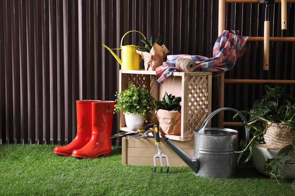 Hermosas plantas, herramientas de jardinería y accesorios en hierba verde cerca de la pared de listones de madera - Foto, imagen