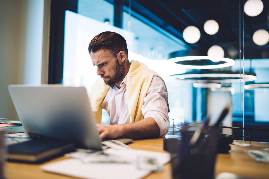 Серйозний концентрований бородатий чоловічий підприємець у формальному одязі, що сидить за дерев'яним столом у сучасному офісі та працює над проектом на ноутбуці
 - Фото, зображення